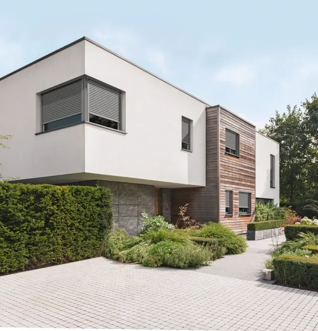 Moderner Alu Rolladen an einem kubischen Einfamilienhaus mit weißer Putzfassade und Holzverkleidung, außenliegend montiert vor rechteckigen Fenstern, geschlossen als Sicht- und Sonnenschutz.