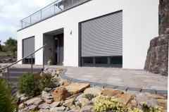 Moderne Hausfassade mit zwei großen geschlossenen Rollläden und gepflastertem Vorplatz mit Steingarten