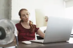 Une femme qui transpire est assise près d'une fenêtre devant un ordinateur portable et un ventilateur, elle utilise une feuille pour s'éventer à cause de la chaleur dans la pièce